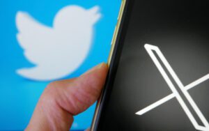 Hand holding phone showing the X logo with Twitter logo in background