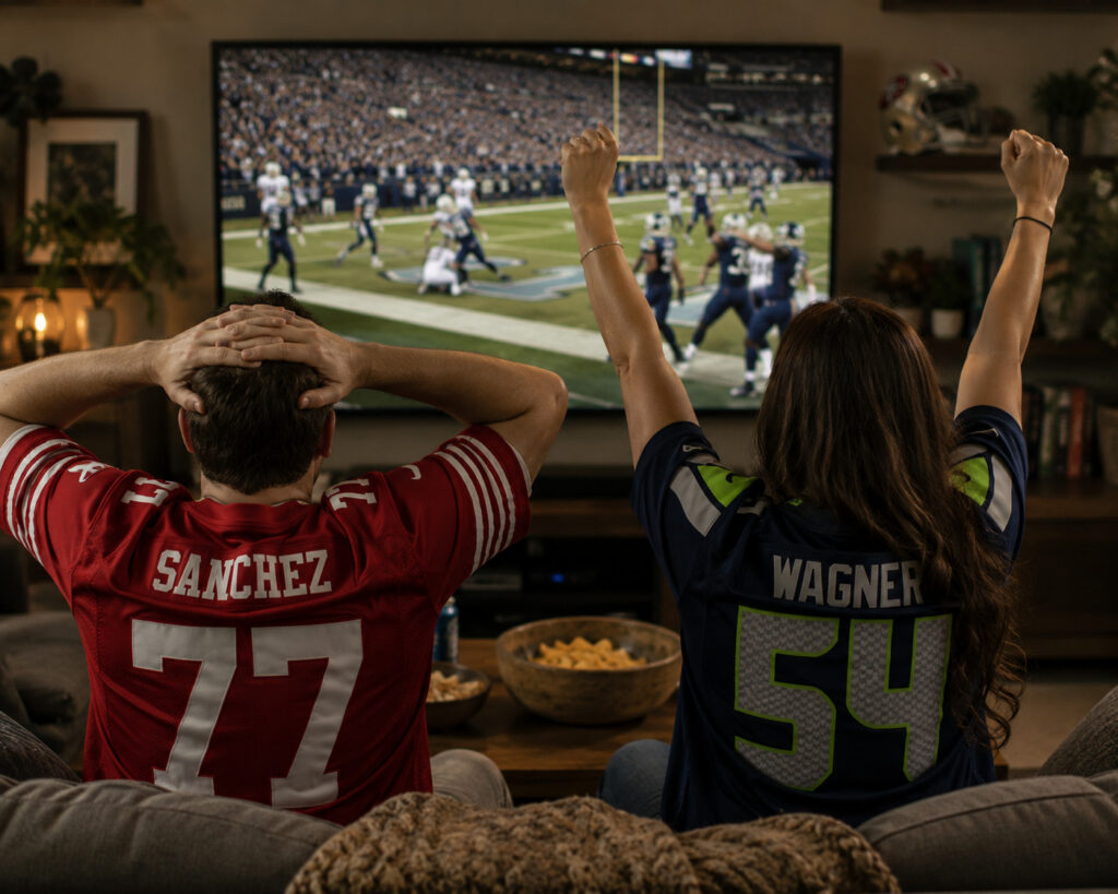 Two sports fans cheering on a couch while watching a football game on television.