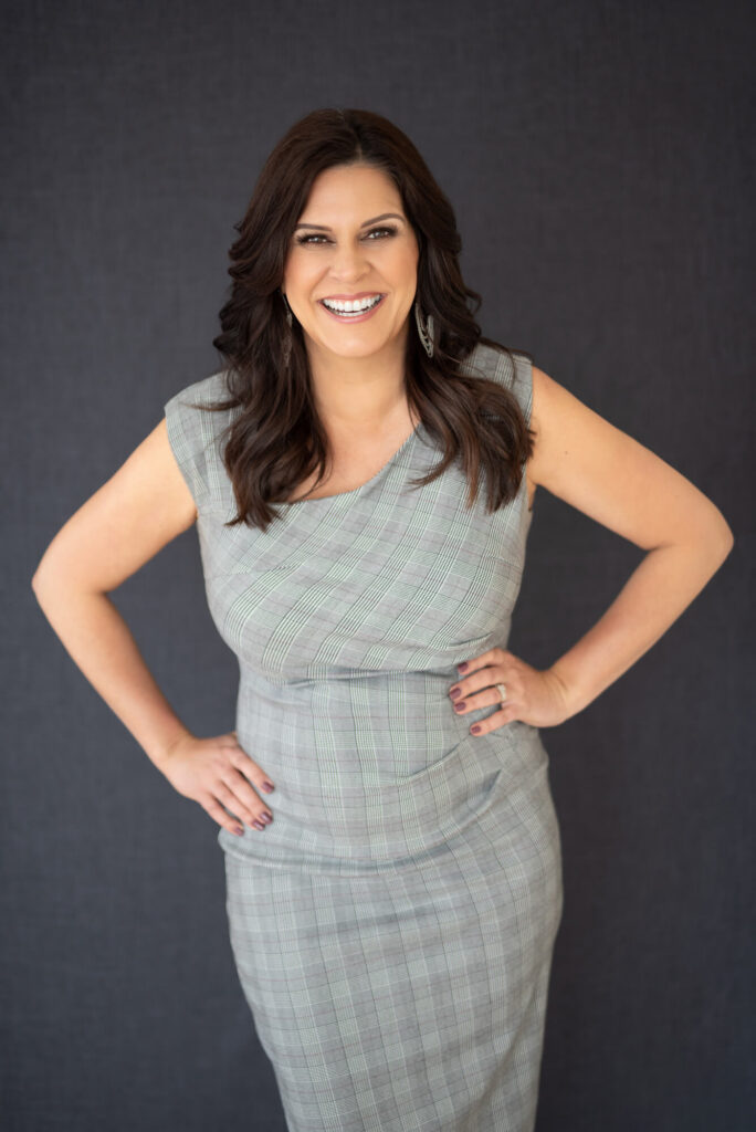 Studio portrait of Viv smiling with her hands on her hips against a dark gray background.
